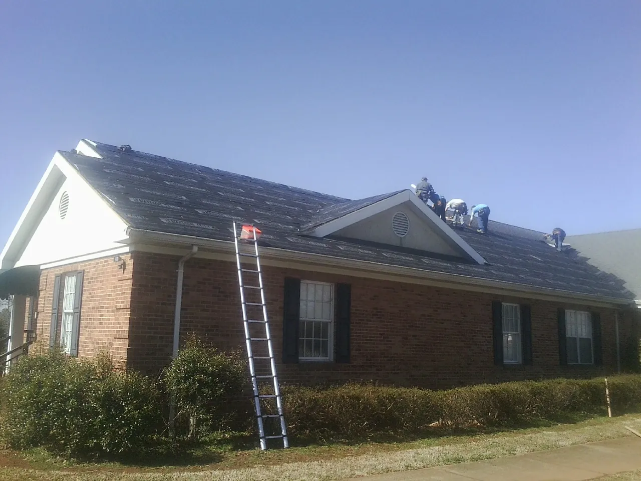 Crew tearing shingles off a brick ranch roof with a ladder set — Emergency Roof Repair work by our crew in Ruthton