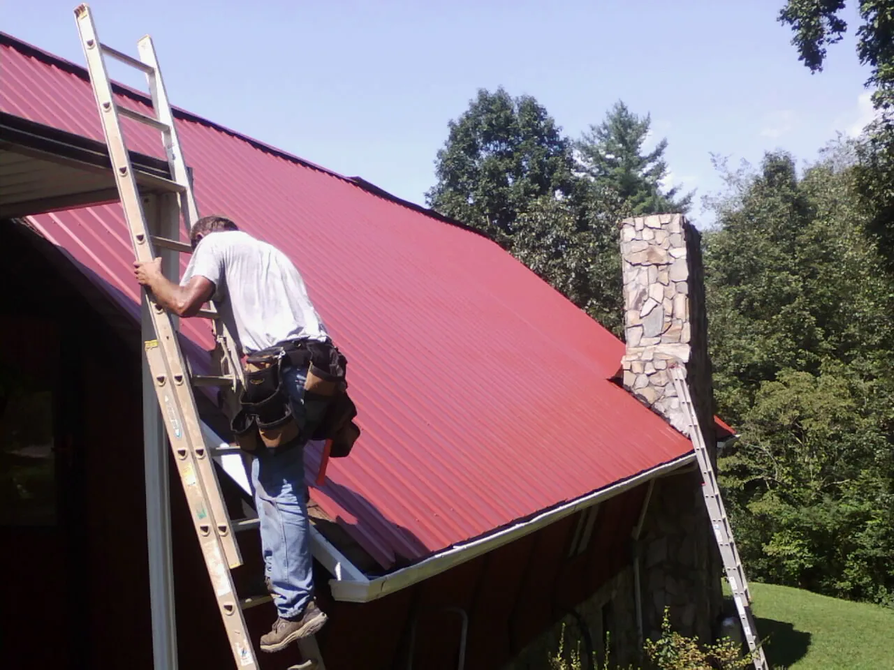 Roofer on a ladder beside a red metal roof and stone chimney — Soffit & Fascia Repair by Roofing Brothers in Ruthton